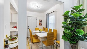 Un comedor moderno con una mesa blanca y sillas amarillas, una planta en maceta, y vistas a una cocina y a un área de estar.