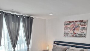 Una cama bien hecha con cubrecama y almohadas de color claro, una mesita de noche con lámpara, y una pared con texto motivacional e ilustración de un árbol.