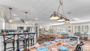 A spacious dining area with a wooden table set for six, surrounded by black chairs. A kitchen with white cabinets and a microwave is visible in the background. - Luna Sea | Anna Maria Beach Home Rental - Steps from the Sea! (Anna Maria)