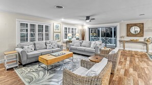 A living room with two grey sofas, a wooden coffee table, wicker furniture, and a large window with blinds. - Luna Sea | Anna Maria Beach Home Rental - Steps from the Sea! (Anna Maria)