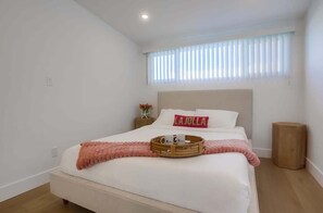 Cama bem arrumada com uma bandeja de objetos, um travesseiro com a inscrição 'LA JOLLA' e uma janela com persianas.