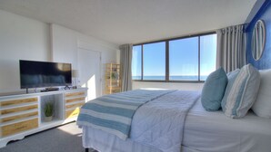 A bedroom with a large bed, a TV on a wooden cabinet, a window with curtains, and a view of the sea.