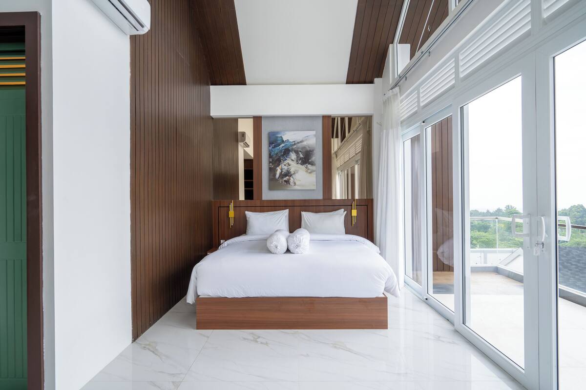 A modern bedroom with a large bed, wooden headboard, and a view of the outdoors through large windows.