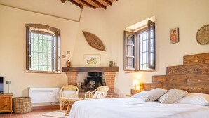 A bedroom with a fireplace, wicker chairs, a bed with striped pillows, and a wooden headboard. - Cosy apartment with pool, A/C, WIFI, TV, patio and panoramic view, close to San Gimignano (Montauto)