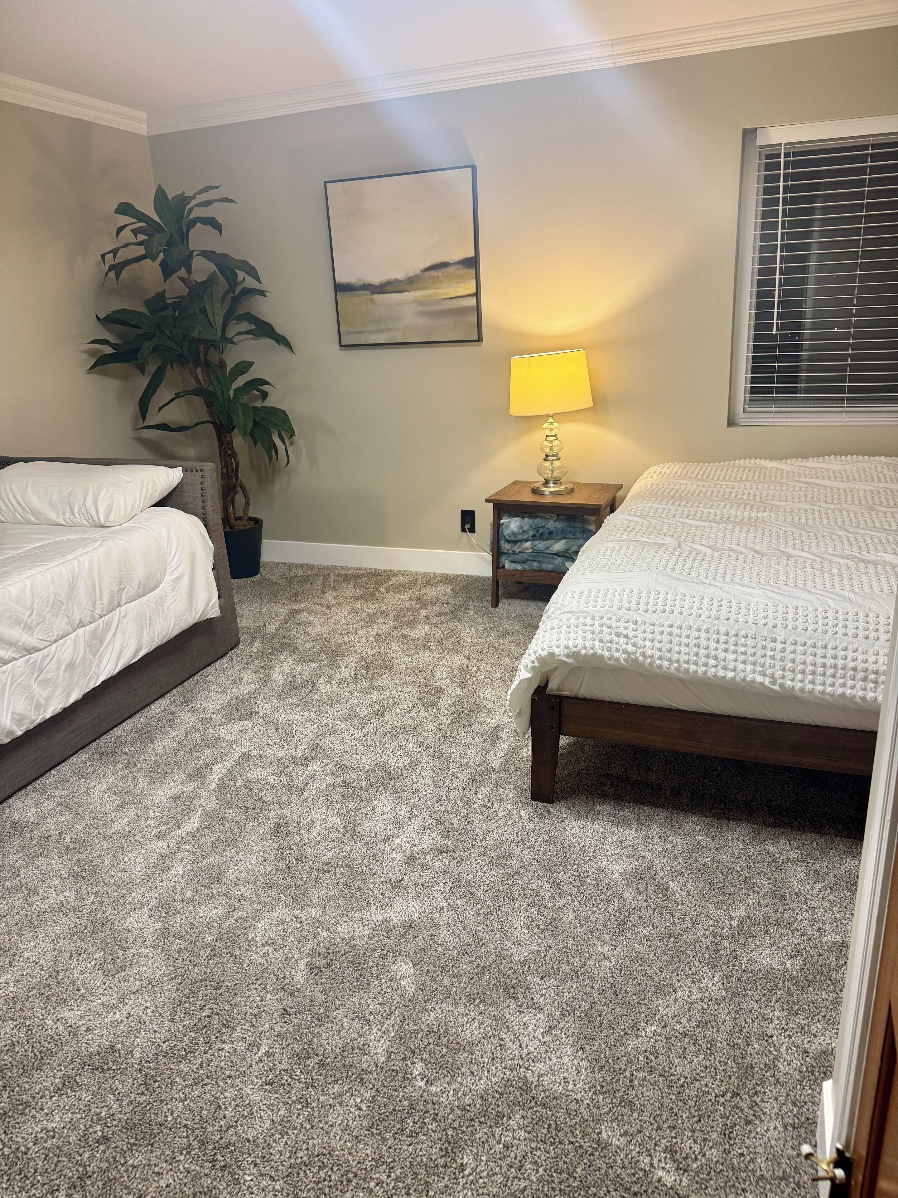 A bedroom with two beds, a large potted plant, a bedside lamp, and a painting on the wall.