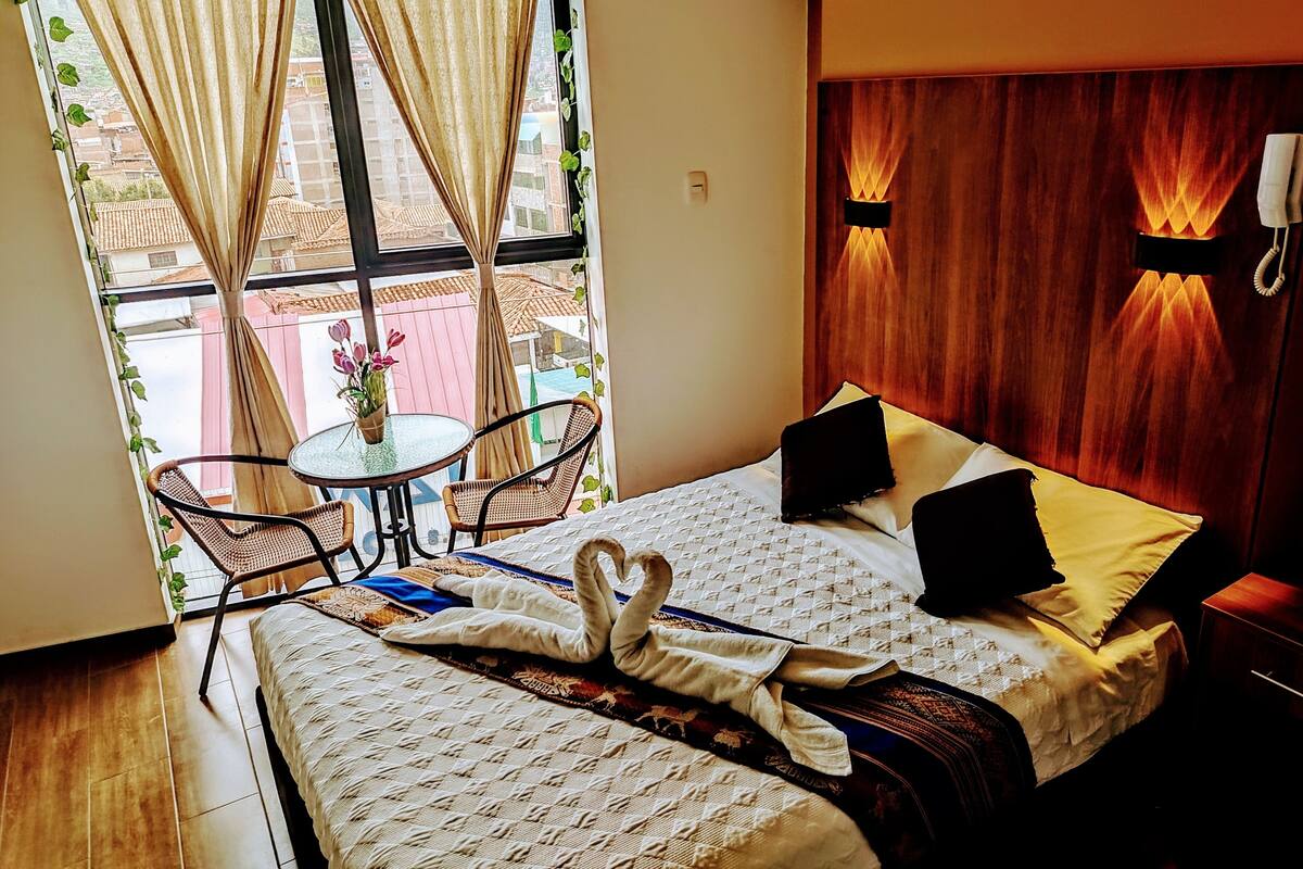 Quarto de hotel com cama, uma pequena mesa com cadeiras, janela com cortinas e uma luminária fixada na parede.