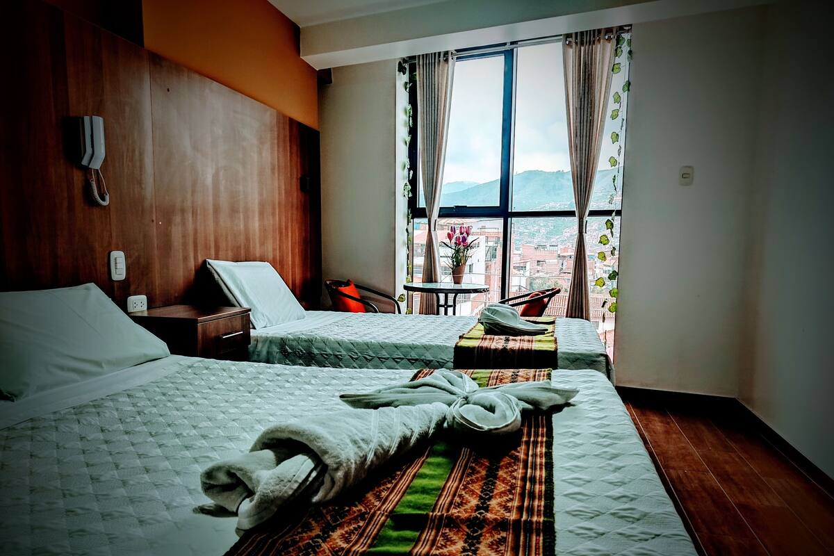 Quarto de hotel com duas camas, uma mesinha com uma planta, uma cadeira e vista para as montanhas pela janela.