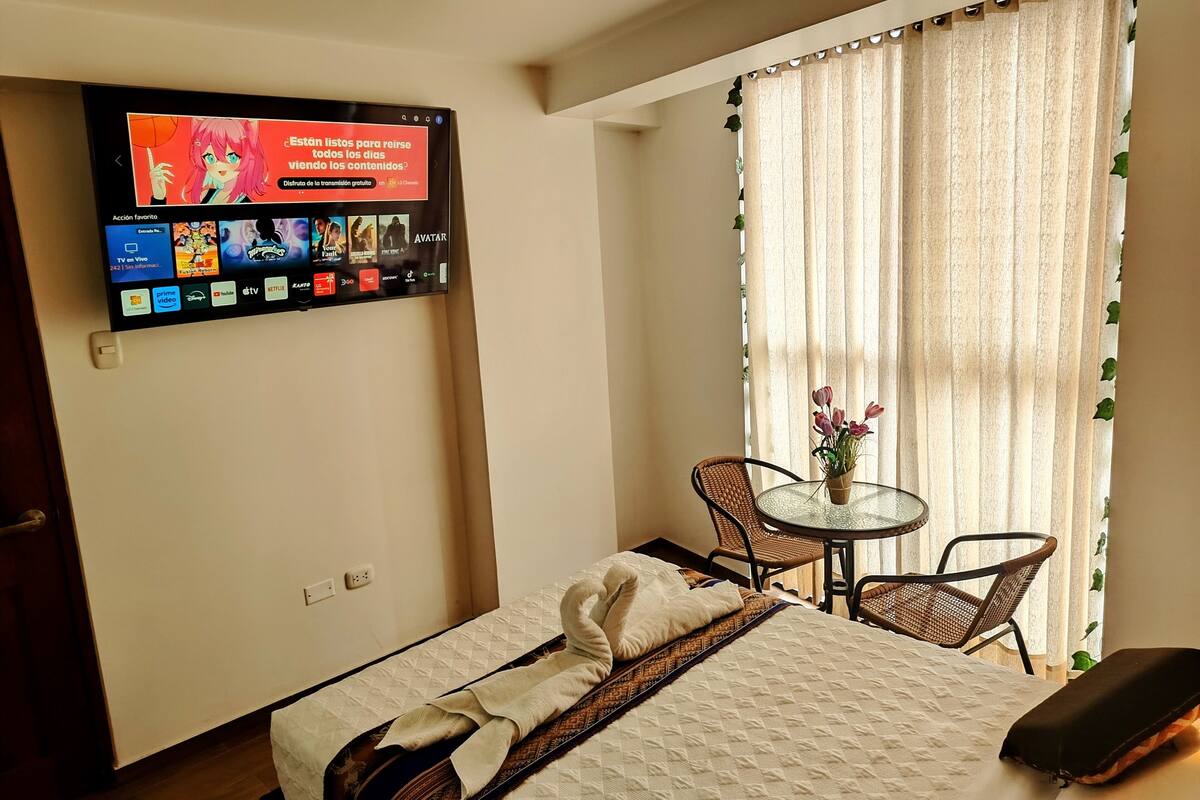 Quarto de hotel com uma cama, televisão fixada na parede exibindo diversos serviços de streaming, uma mesa redonda pequena com uma cadeira, e uma janela com cortinas.