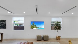A modern living room with a large flat-screen TV, two ottomans, a brown leather sofa, a small round table, and a potted plant. - Up to 12 guest Modern Villa near Wynwood! (Miami)