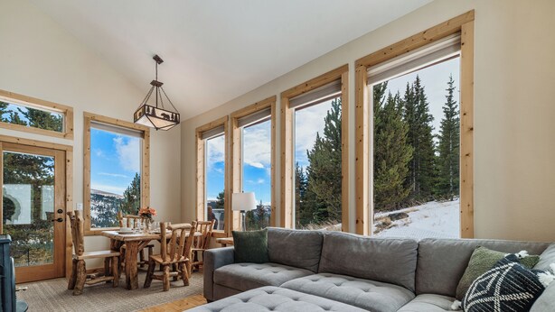 Un cuarto acogedor con un amplio sofá seccional gris, una mesa de comedor de madera y sillas. Amplias ventanas ofrecen vistas a árboles y un paisaje nevado.