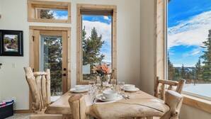 Una mesa de comedor de madera dispuesta con platos, tazones y vasos blancos, rodeada de sillas de madera, con vista a árboles y montañas a través de ventanas amplias.