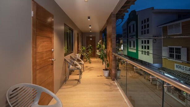 Een gang met houten deuren, een zithoek met stoelen, potplanten en een uitzicht op gebouwen achter een glazen balustrade.