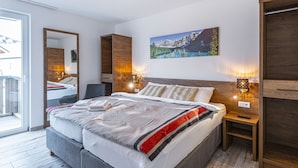 Een moderne slaapkamer met een groot bed, een houten hoofdbord, twee nachtkastjes met lampen, een grote spiegel en een schilderij van een berglandschap.