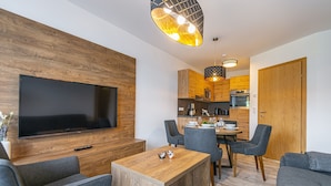 Een moderne woonkamer met een flatscreen televisie, grijze banken, een houten salontafel en een eethoek met een tafel en stoelen.