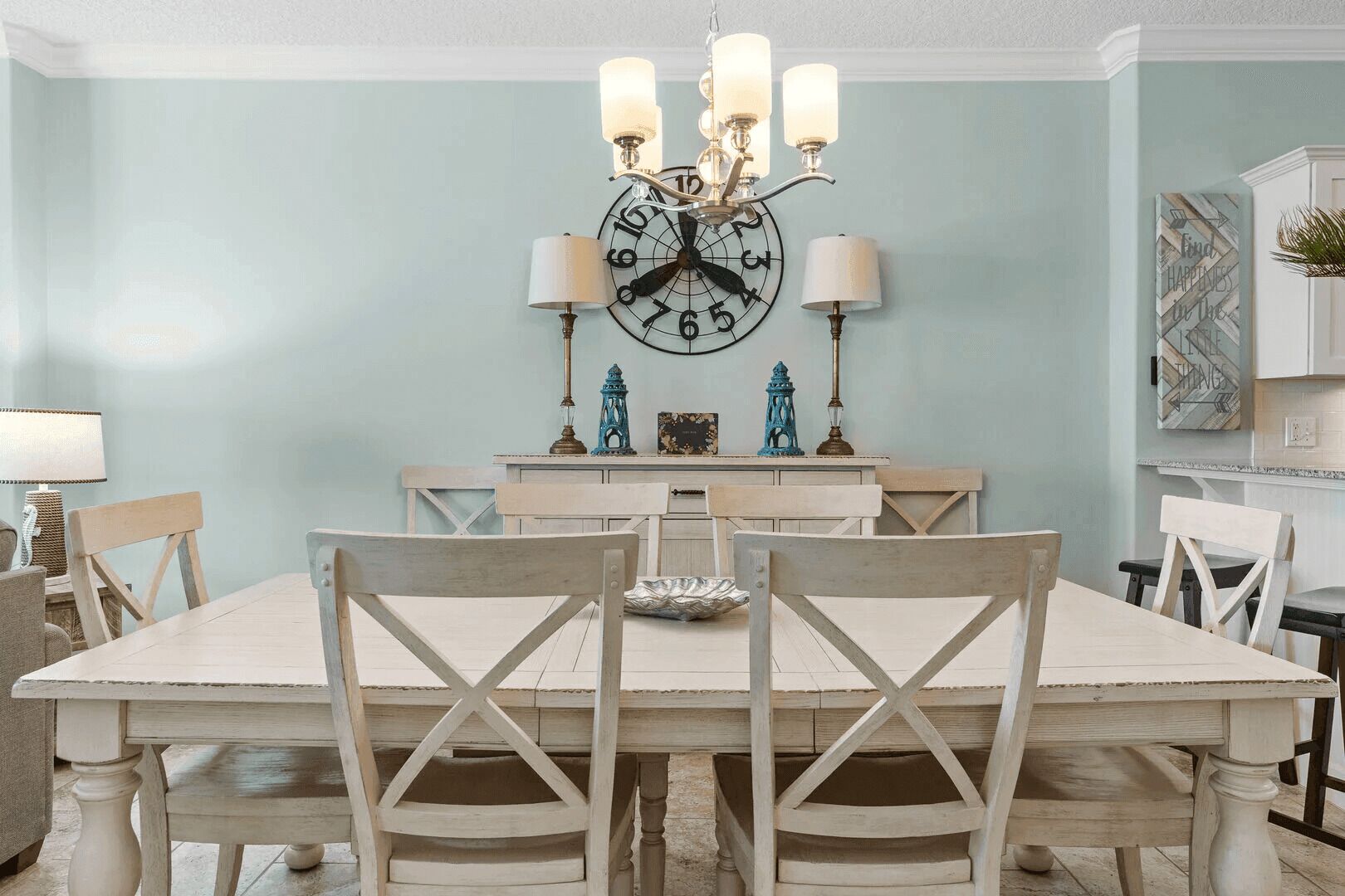 Une salle à manger avec une grande table en bois, des chaises blanches, un lustre et une horloge murale.