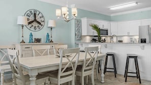 Un comedor con mesa y sillas de madera, un reloj de pared y una cocina con armarios blancos y un microondas.