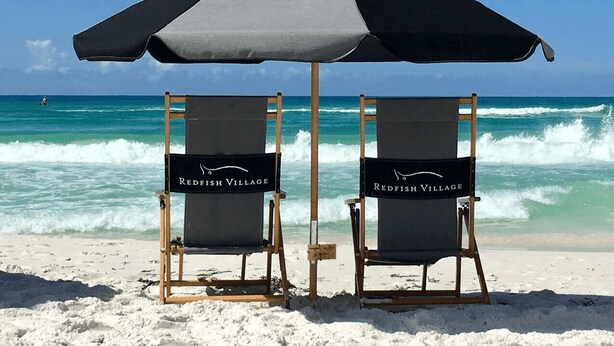 Two beach chairs labeled "Redfish Village" under a large umbrella on a sandy beach with the ocean in the background. - Hello 30a! by Avantstay Private Beach Access + Shared Pool (Santa Rosa Beach)