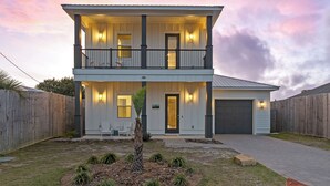 A two-story house with a front porch, a garage, and a small garden with plants. - Wanderlust by Avantstay Walk to Beach + Pool New Construction (Panama City Beach)