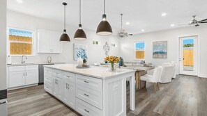 A modern kitchen with white cabinetry, a central island, pendant lighting, and a dining area with white chairs. - Wanderlust by Avantstay Walk to Beach + Pool New Construction (Panama City Beach)