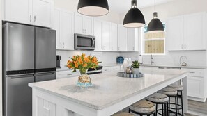 A modern kitchen with white cabinets, a stainless steel refrigerator, a microwave, and a large island with bar stools. - Wanderlust by Avantstay Walk to Beach + Pool New Construction (Panama City Beach)