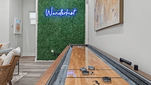 A shuffleboard table with numbered pucks, a neon sign reading 'Wanderlust,' and a green wall with foliage.
