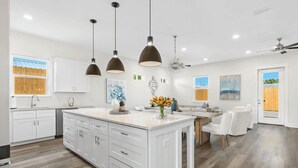 A modern kitchen with white cabinetry, a central island, pendant lighting, and a dining area with a white table and chairs. - Wanderlust by Avantstay Walk to Beach + Pool New Construction (Panama City Beach)