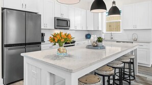 A modern kitchen with white cabinetry, stainless steel appliances, a large island with stools, and pendant lighting. - Wanderlust by Avantstay Walk to Beach + Pool New Construction (Panama City Beach)
