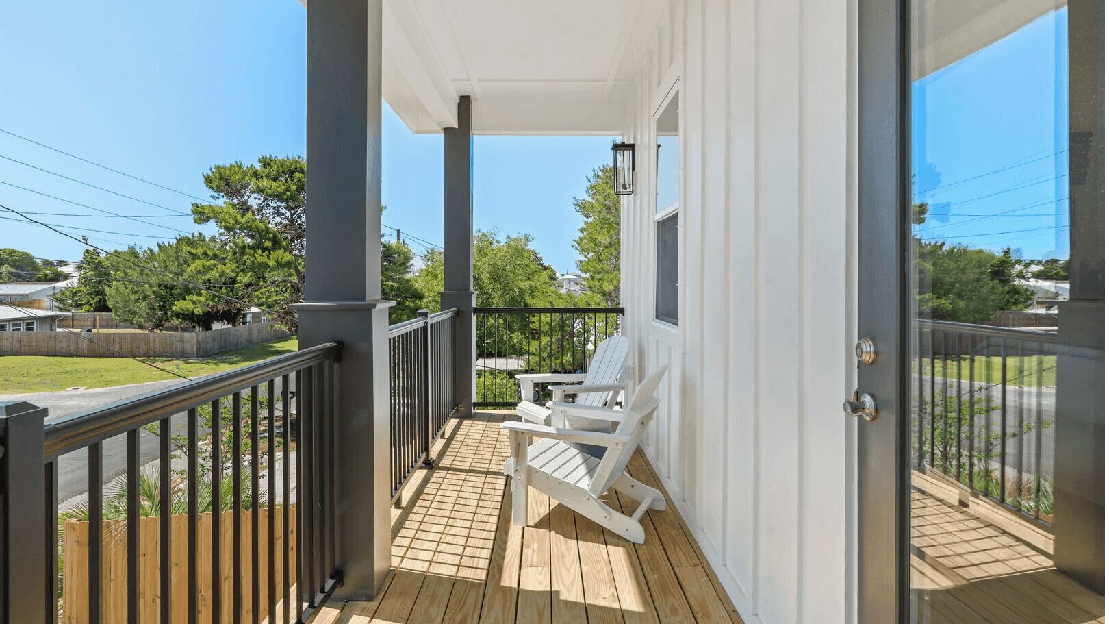 A covered porch with a view of a street and trees, two white Adirondack chairs, and a wooden deck.