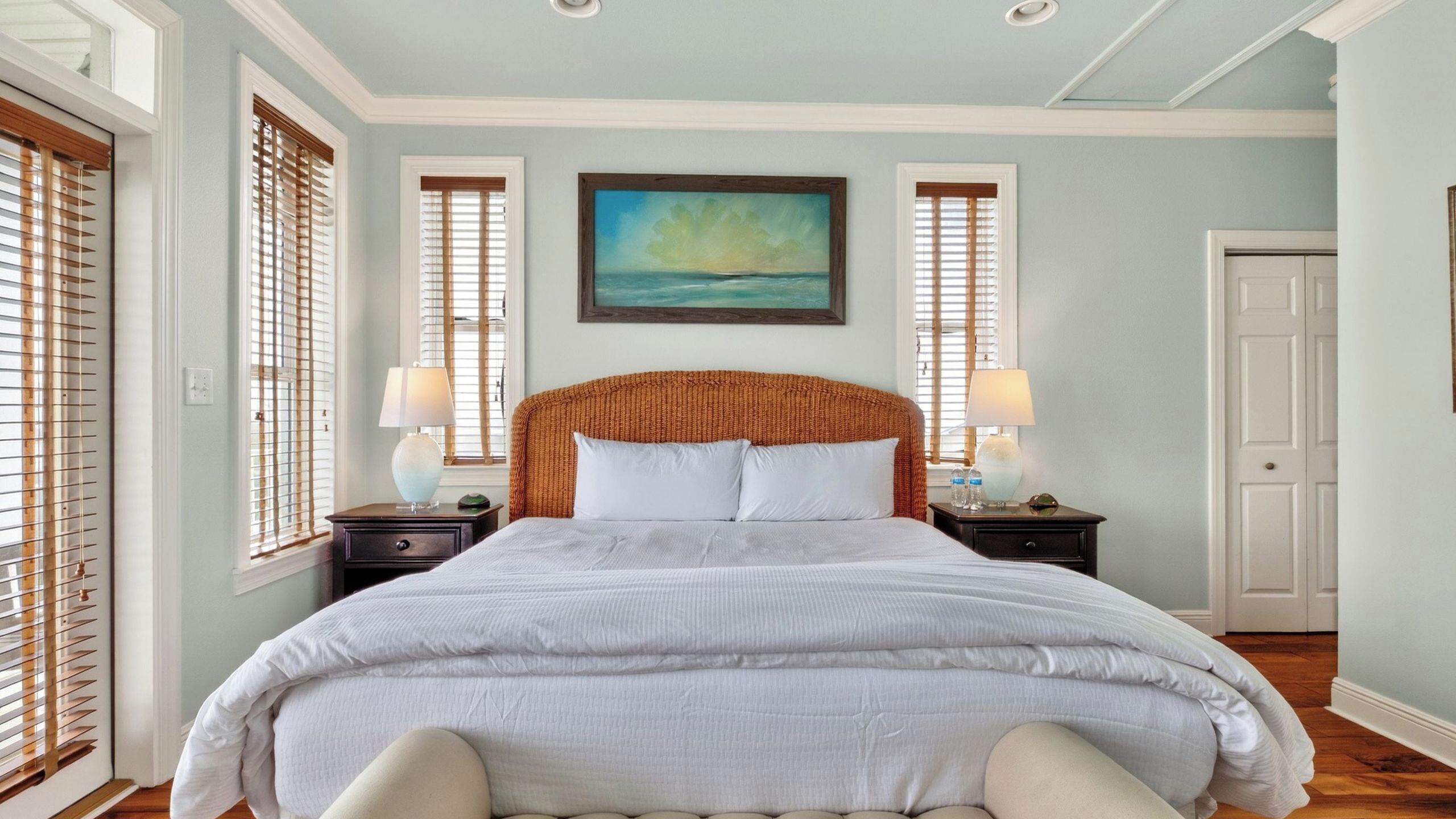 A bedroom with a large bed, two bedside tables with lamps, a painting above the bed, and a door to the right.
