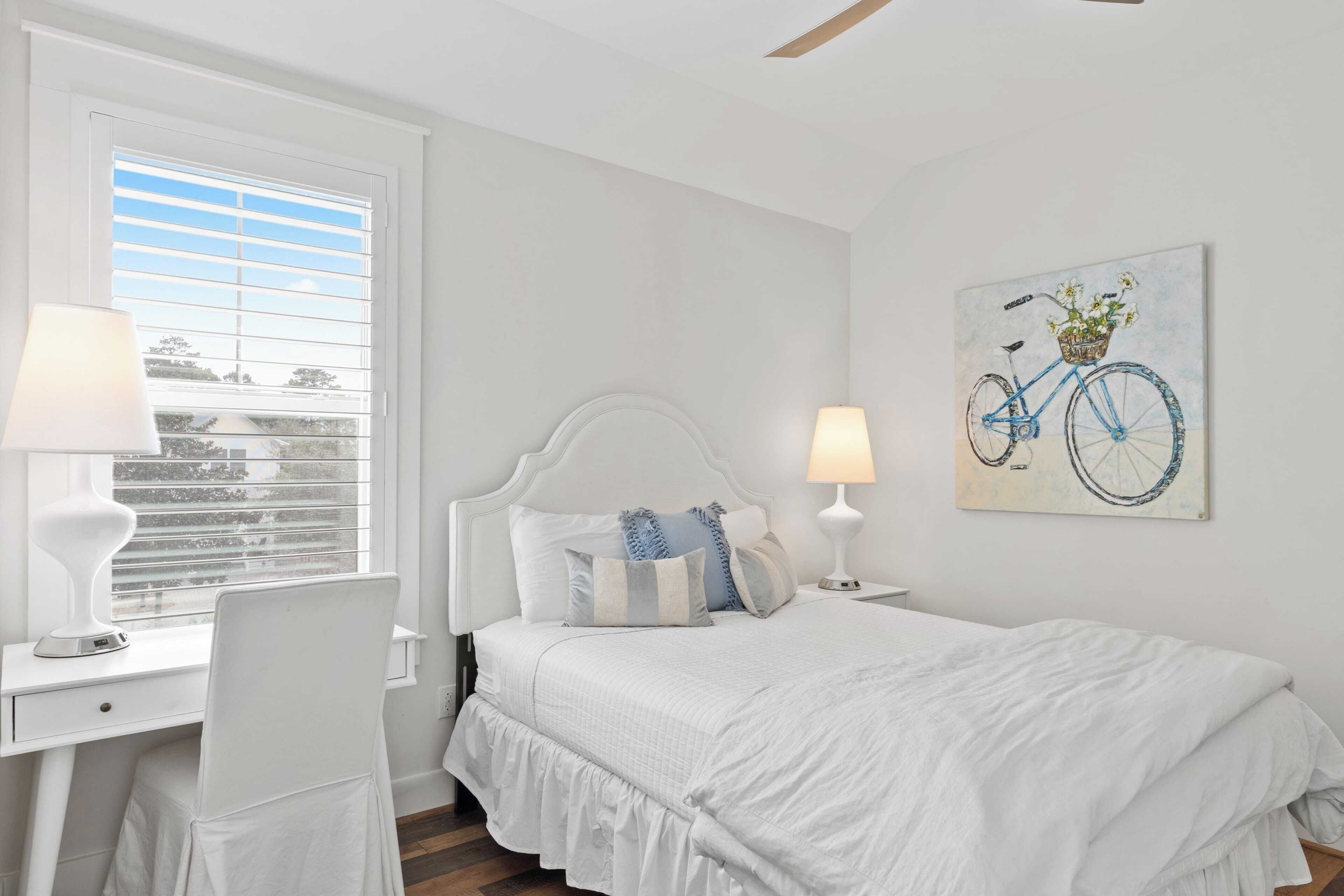 A bedroom with a white bed, two bedside lamps, a desk with a chair, and a window with blinds.