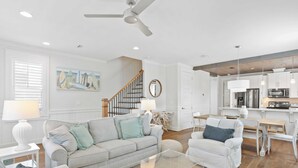 Ein modernes Wohnzimmer mit einem grauen Sofa, weißen Sesseln und einem Glastisch. Eine Treppe mit Holzwand bietet Zugang zu einer Küchenzeile mit weißen Schränken und einem Edelstahl-Kühlschrank.