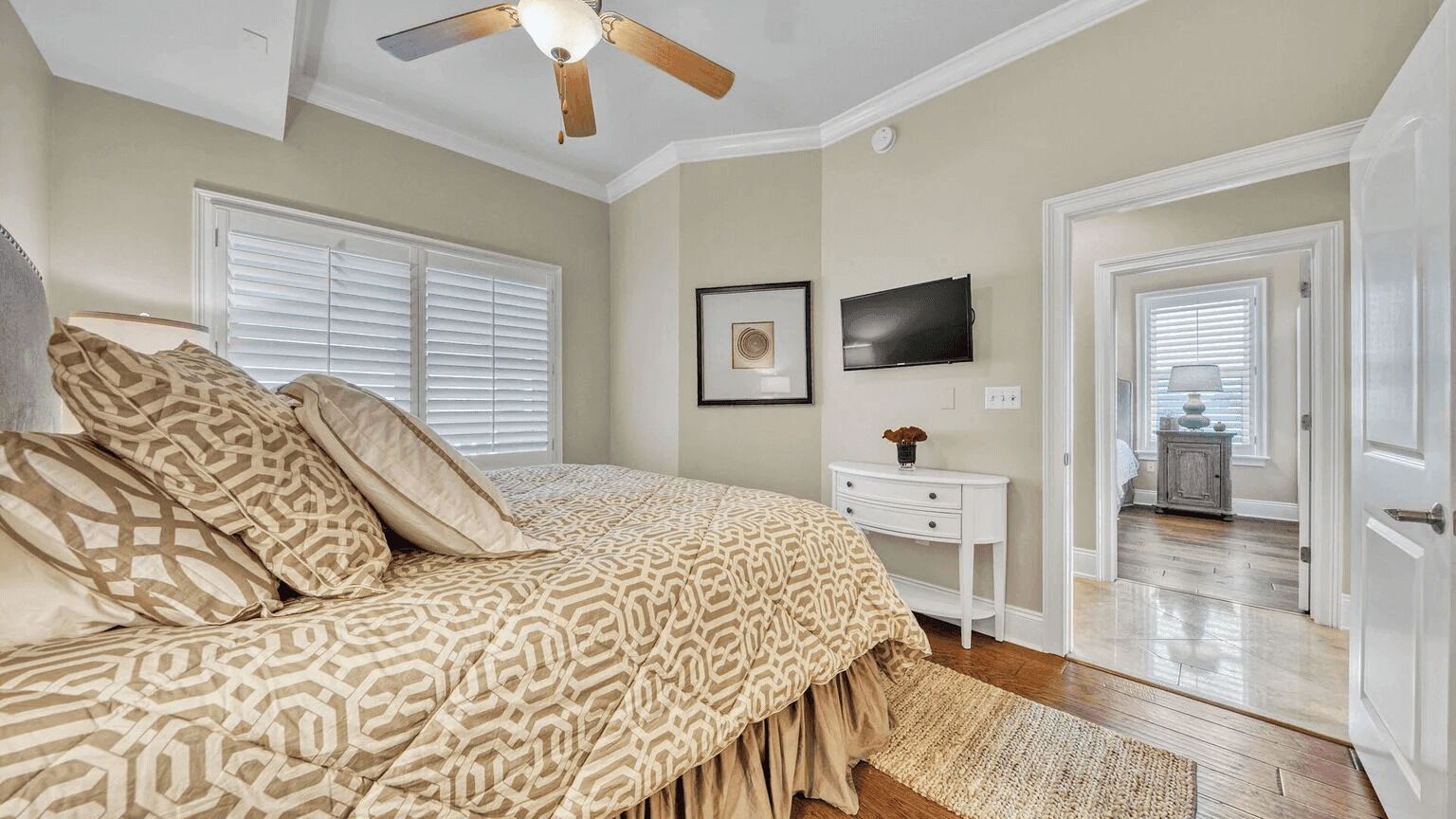 A bedroom with a bed, a ceiling fan, a TV, a bedside table, and a door leading to another room.