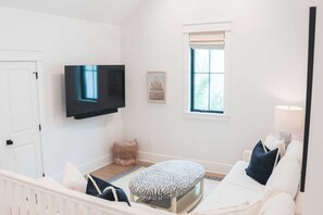 A modern, well-lit room with a white sofa, a zebra-patterned ottoman, a flat-screen TV, a wicker basket, and a window with blinds. - Shellom by Avantstay Newly Built, Pool Access (Santa Rosa Beach)