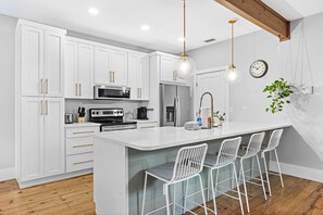A modern kitchen with white cabinets, a central island with bar stools, stainless steel appliances, and pendant lighting. - Bayou Bungalow by Avantstay Walk to Boat Ramp & Bay Private Yard (Santa Rosa Beach)