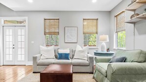 A living room with a grey sofa, a green armchair, and a brown ottoman. There are two windows with woven blinds, a white lamp on a side table, and a framed abstract art piece on the wall. - Bayou Bungalow by Avantstay Walk to Boat Ramp & Bay Private Yard (Santa Rosa Beach)