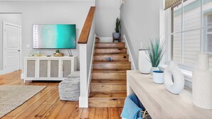 A staircase with wooden steps leading to a landing, a white console table with decorative items, a flat-screen TV on the wall, and a window with blinds. - Bayou Bungalow by Avantstay Walk to Boat Ramp & Bay Private Yard (Santa Rosa Beach)