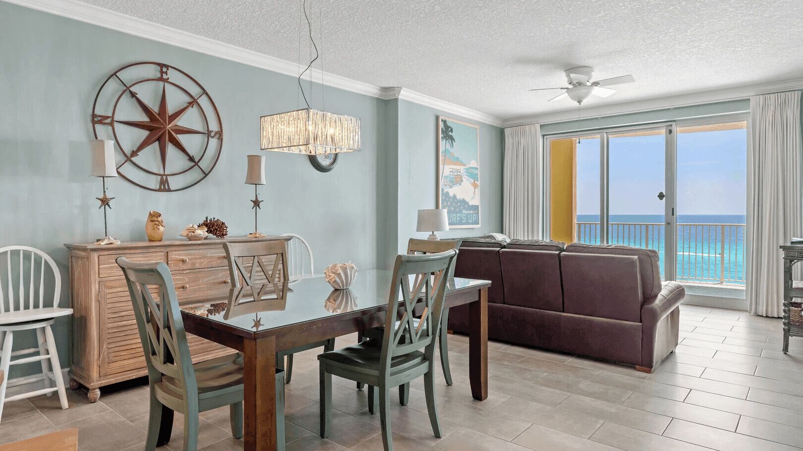 A living room with a dining area, a large window with a view of the ocean, a ceiling fan, and a wall-mounted compass.