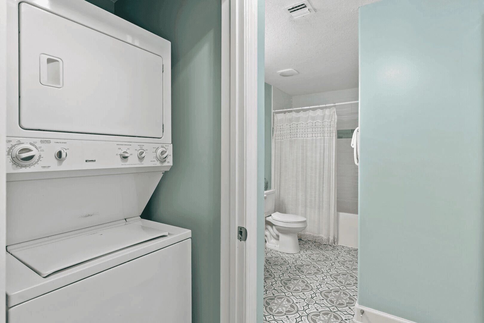 A laundry area with a washing machine and dryer, a bathroom with a toilet and shower curtain, and patterned floor tiles.