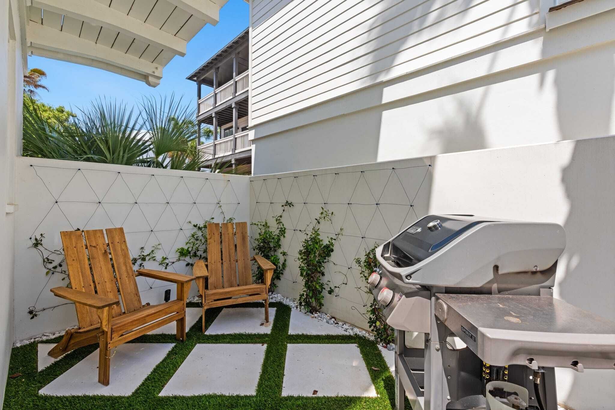 A patio area with two wooden Adirondack chairs, a barbecue grill, and a white fence with climbing plants.