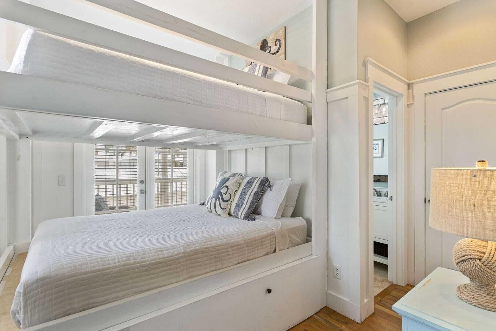 Une chambre avec un lit superposé, décorée dans des tons de blanc, comprenant une table de chevet avec une lampe et une fenêtre avec des stores.