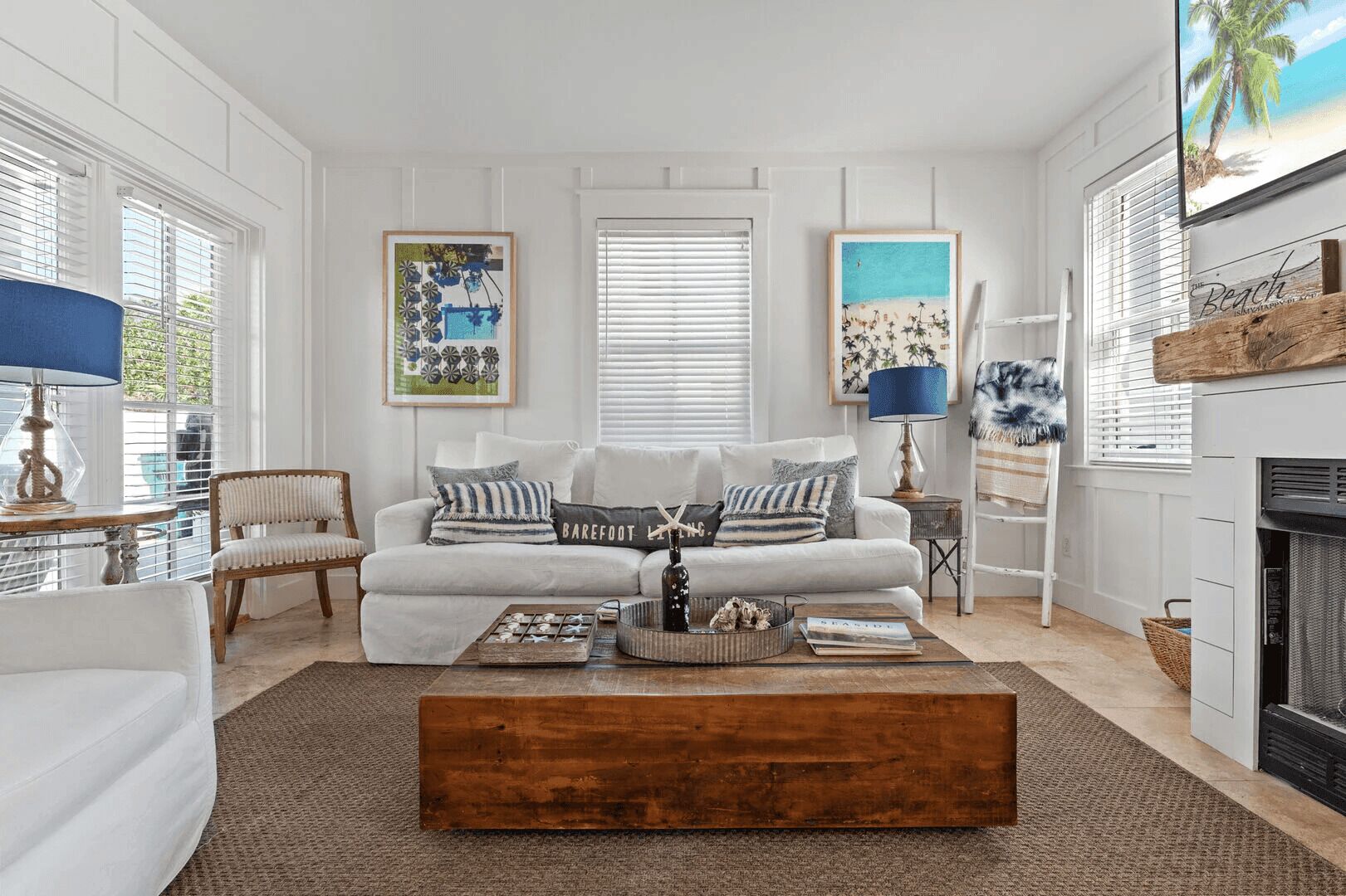 A living room with a white sofa, a wooden coffee table, and a fireplace. There are framed pictures on the wall and a lamp on a side table.
