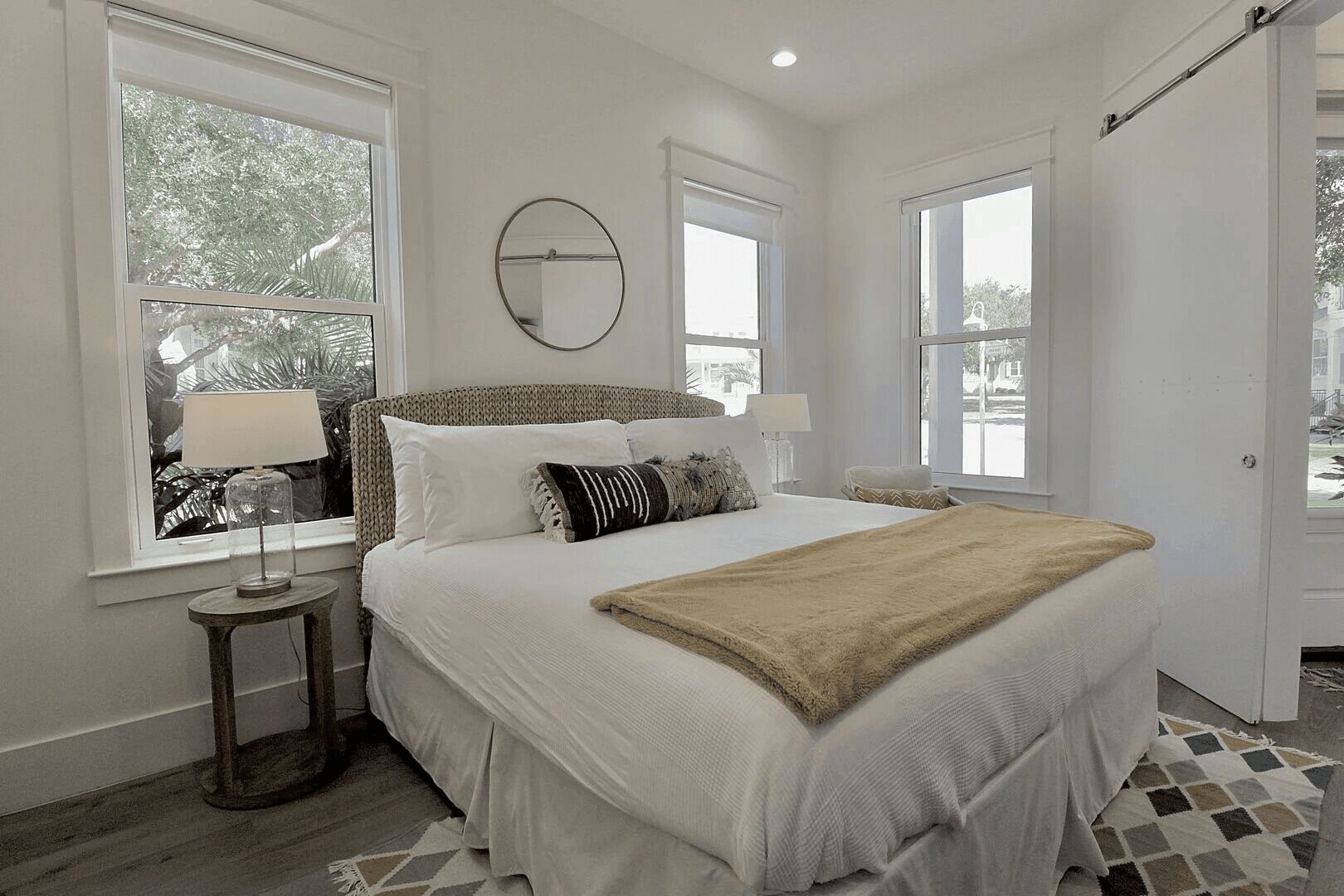 A bedroom with a large bed, two bedside tables, a round mirror, and multiple windows.