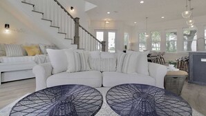 A spacious living room with a white sofa, two black wire-framed tables, a staircase, and a kitchen area with bar stools. - Jessa Blu by Avantstay Walk to Beach Pool + Gym (Santa Rosa Beach)