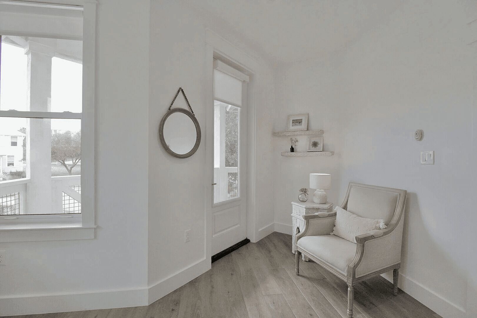 A spacious room with a large window, a white chair, a small table with a lamp, and a decorative mirror on the wall.