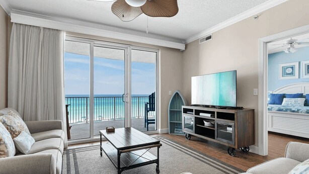 A spacious living room with a large sliding glass door leading to a balcony with a view of the ocean. There is a flat-screen TV on a wooden stand, a sofa, and a coffee table. - Emerald Isle Resort 405 by Avantstay Beachfront, Beach Chairs + Pool Access (Panama City Beach)