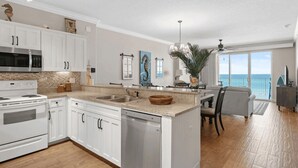 A spacious kitchen with white cabinetry, a large island with a sink, and a view of the ocean through a sliding glass door. - Emerald Isle Resort 405 by Avantstay Beachfront, Beach Chairs + Pool Access (Panama City Beach)