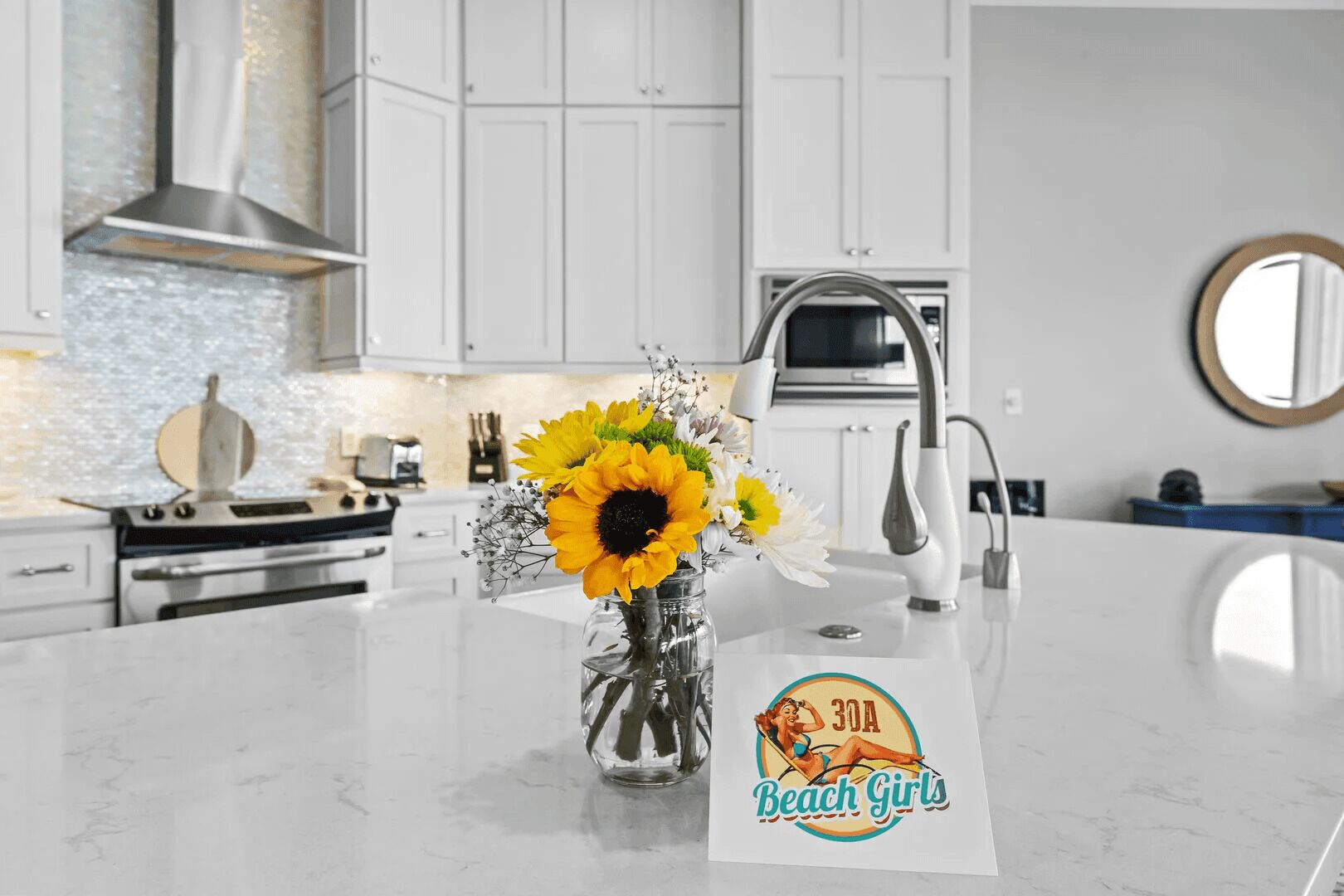 A modern kitchen with white cabinets, a stainless steel range hood, a built-in oven, and a large island with a vase of sunflowers.