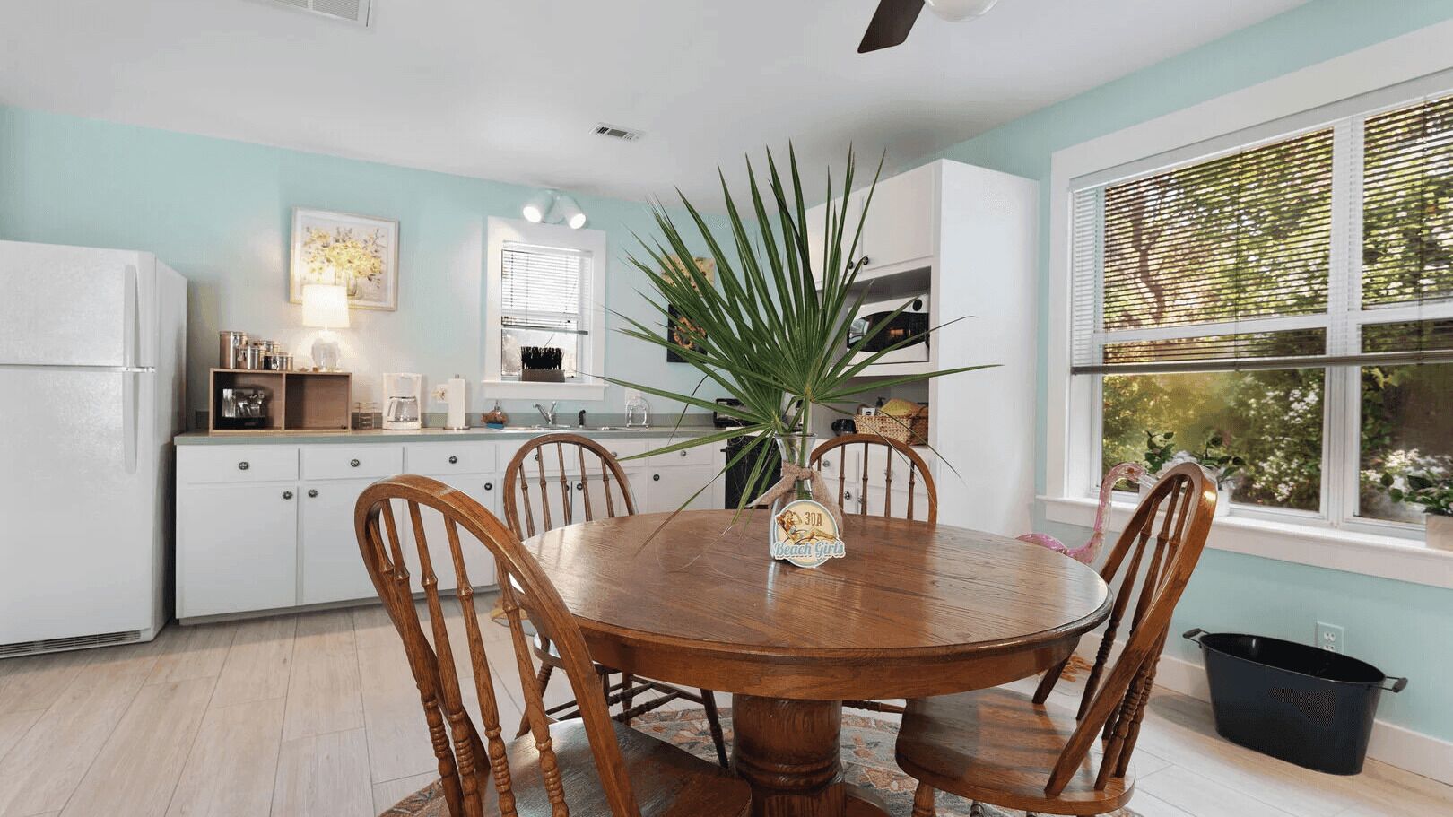 A kitchen with a round wooden table, white cabinets, and a large window with blinds.