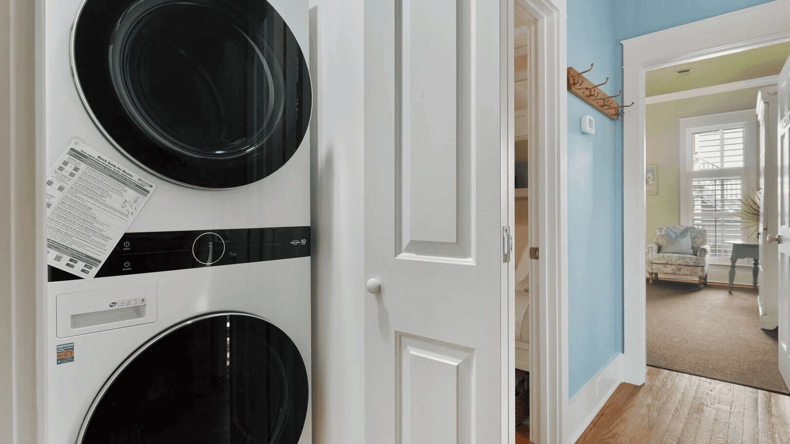 A laundry room with two washing machines, a door leading to a living area with a sofa and a window.