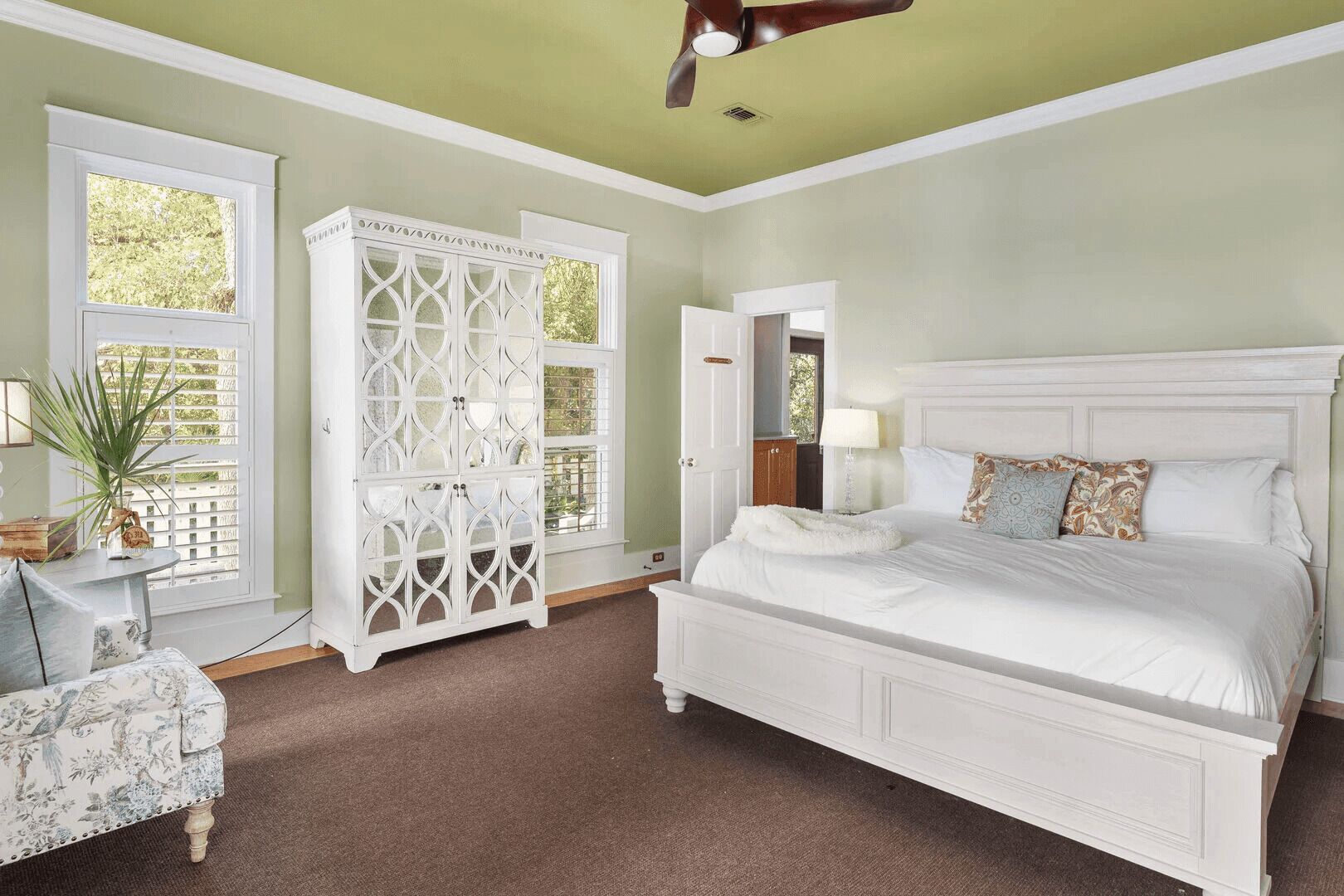 A bedroom with a large bed, a white wardrobe, a small table with a lamp, and a chair.